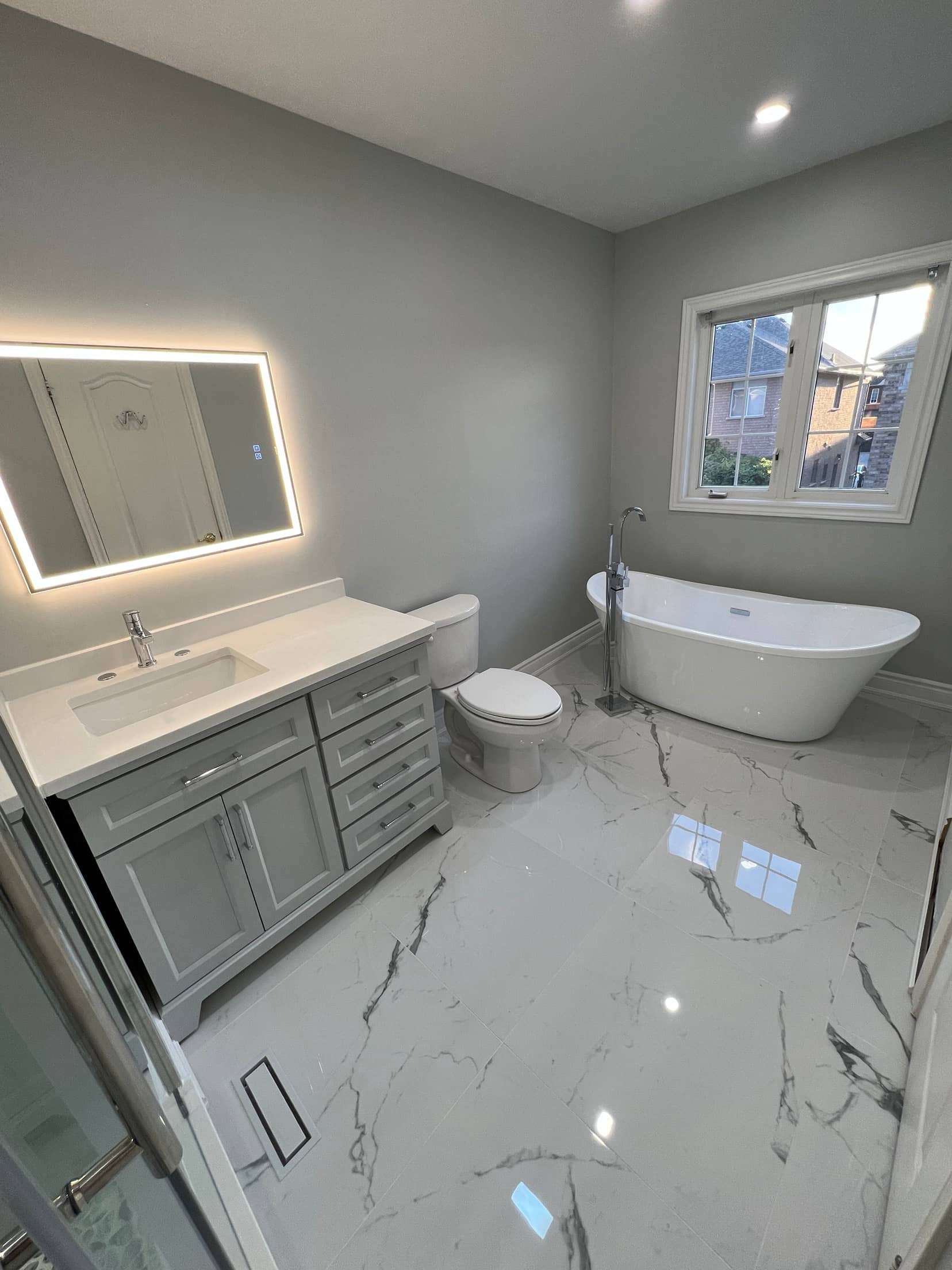 Grey shaker vanity with a backlit mirror, polished marble look floor tile and a freestanding tub by the window