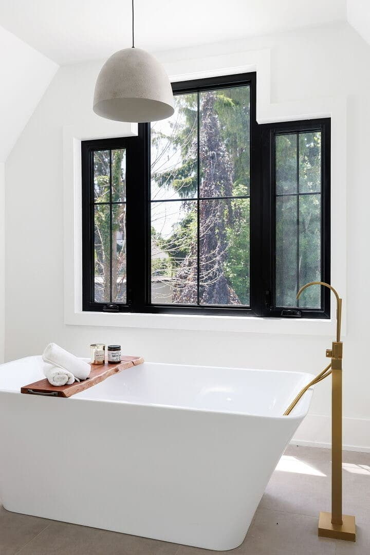 Freestanding tub beneath black framed windows, with a brushed gold floor mounted filler and a timber bath caddy