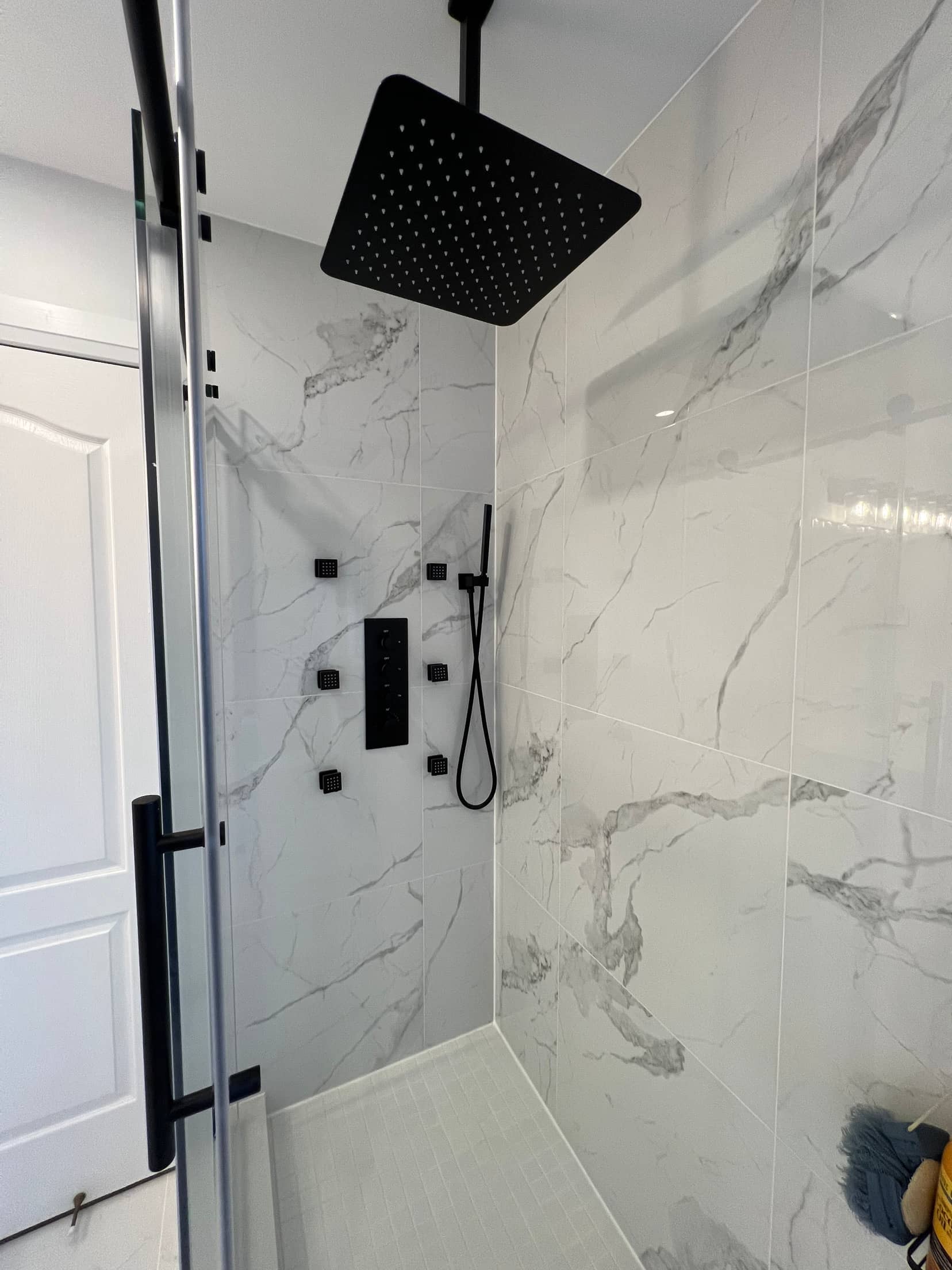Marble tiled shower with a black rainfall head, body jets and a thermostatic valve behind frameless glass